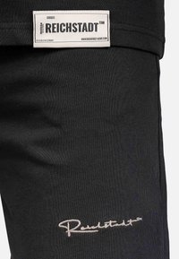 Zwarte stoffen broek met een wit merklabel en een in zilver geborduurd "Reichstadt"-logo. Soepele textuur met een aansluitend ontwerp.