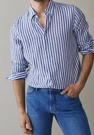 Mann mit hochgekrempelten Ärmeln, trägt ein blau-weiß gestreiftes Hemd mit Knöpfen, in blaue Jeans gesteckt, Hand in der Tasche.