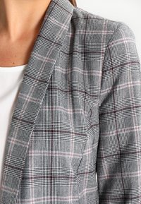 Šedý kostkovaný blazer s texturovanou tkaninou, tenkými červenými a bílými pruhy, s klopovým límcem a pevným ramenním prostorem.