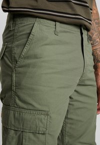 Olivengroene cargobroek van structuurstof, met zijzakken en een rechte snit. Subtiel ruitpatroon zichtbaar op het materiaal.