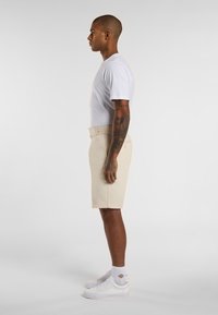 Weißes T-Shirt mit Rundhalsausschnitt, beige Shorts und weiße Turnschuhe. Tätowierte Arme sind sichtbar. Shorts mit Gürtelschlaufen, taillierte Passform, glatte Stofftextur.
