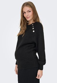 Nosečnica, oblečena v črni pulover s srebrnimi gumbi na rami in črno plisirano krilo, stoji ob svetlem ozadju.