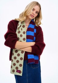 Pletená šatka so zeleným pozadím s hnedými bodkami a modro-vínovo pruhovanou šatkou, držaná v neformálnej póze.