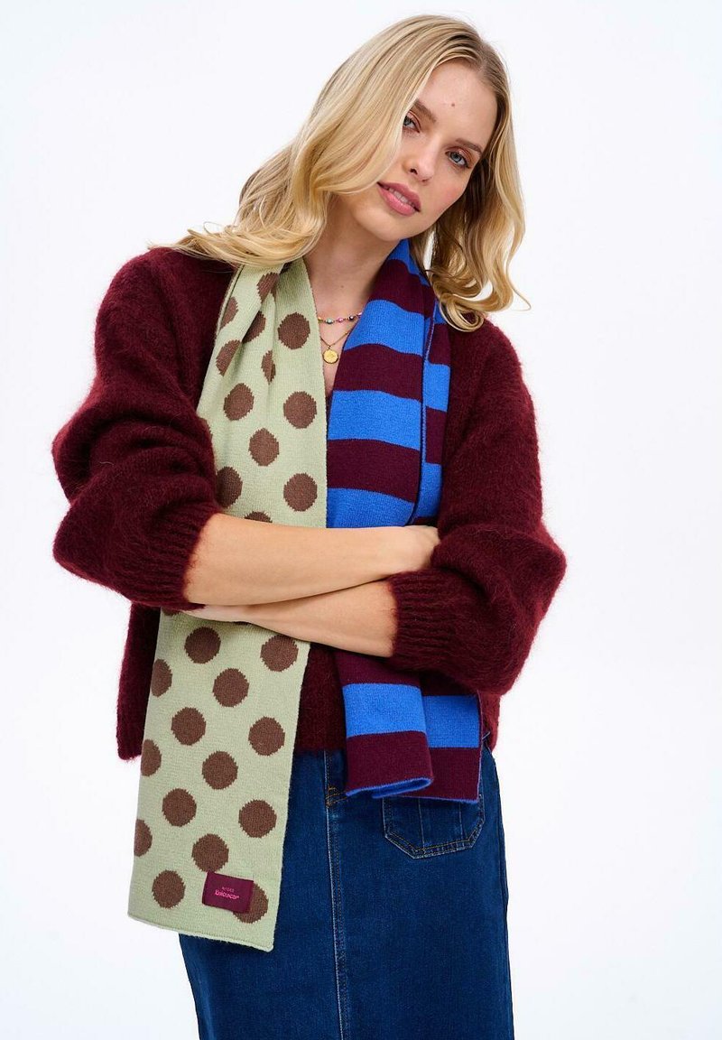 Pletená šatka so zeleným pozadím s hnedými bodkami a modro-vínovo pruhovanou šatkou, držaná v neformálnej póze.
