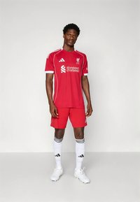 Rdeč nogometni komplet Liverpool FC z belimi poudarki, z logotipom, blagovno znamko "Standard Chartered" in belimi nogavicami s črnimi črtami.