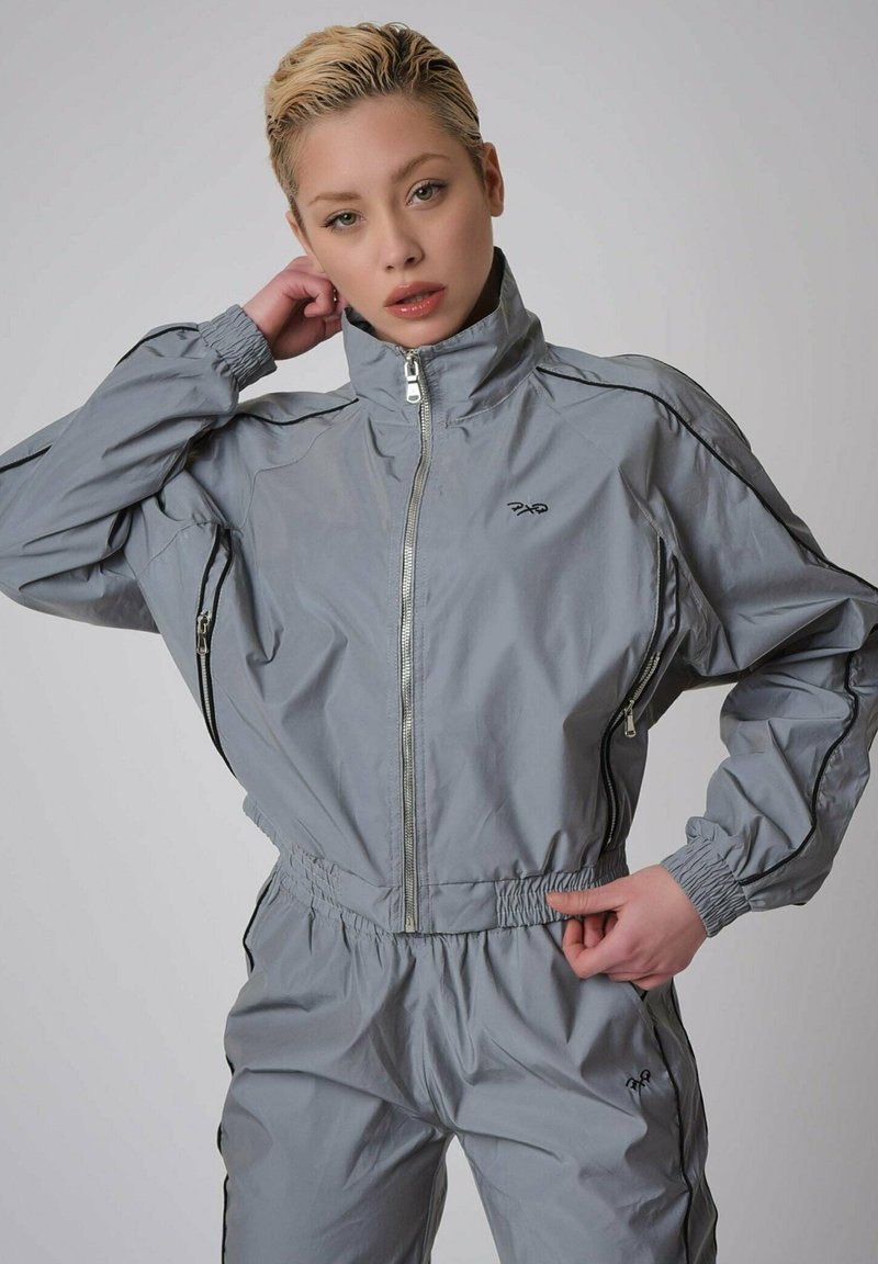Survêtements gris en matériau léger et brillant. Caractéristiques d'un col montant, de poches zippées, de poignets élastiques et d'une taille élastique ajustée.