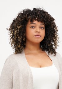 Light beige cardigan with a soft texture, open front, and wide sleeves, worn over a white top. Curly hair with dark and light highlights.