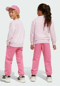 Ensemble sweat-shirt et pantalon de jogging rose avec des poignets élastiques, une coupe décontractée, et une casquette rose assortie, accompagné de baskets blanches et noires.
