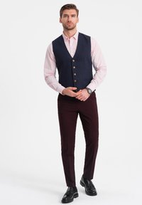 Gilet a quadri blu navy con cinque bottoni sopra una camicia rosa, abbinato a pantaloni bordeaux e scarpe nere, creando un outfit formale.