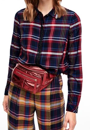 Camicia a quadri in blu scuro, rosso e grigio con chiusura a bottoni. Pantaloni a quadri marroni e gialli. Borsa a tracolla rossa metallica con diverse cerniere.
