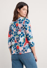 Fløjl med flerfarvet blomstermønster og 3/4 ærmer, med levende blå, røde og grønne nuancer mod en struktureret baggrund.