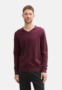 Suéter burdeos de cuello en v, de mangas largas, textura suave, con cuello y dobladillo acanalados, llevado sobre pantalones negros, con un pequeño detalle de logo en el pecho.