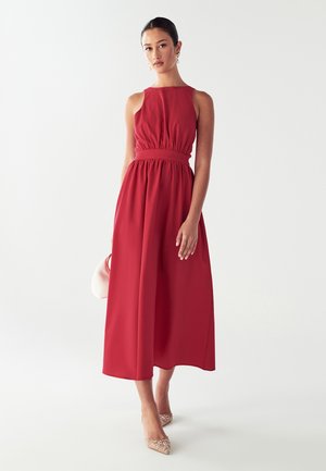 Abito maxi rosso senza maniche con vita arricciata, gonna fluida e texture di tessuto liscia. Abbinato a tacchi chiari e decorati.