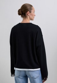 Femme aux cheveux bruns en chignon, portant un pull noir avec des poignets en dentelle et un jean bleu clair, vue de dos sur un fond neutre.