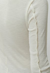 Lichtbeige lange mouw top met een textuur en ribbelpatroon; heeft een golvende naad langs de arm voor extra detail.