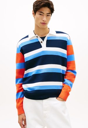 Jonge man draagt een rugbyshirt met lange mouwen in horizontale strepen van marineblauw, blauw, wit en oranje, gecombineerd met een witte broek, handen in de zakken.