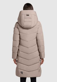 Persoon in een lange, gewatteerde beige winterjas met een grote capuchon, met horizontale en chevron-stiksel, gezien vanaf de achterkant.