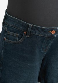 Vaqueros de denim azul oscuro con un diseño de cinco bolsillos, acentos de herrajes de cobre y costuras en contraste a lo largo de la cinturilla y los bolsillos.