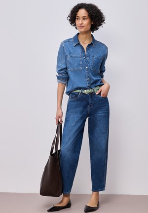 Donna in piedi con camicia di jeans e jeans, che tiene una borsa a tracolla marrone scuro, indossa ballerine nere e una cintura intrecciata verde.