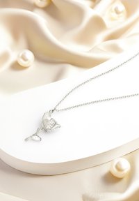 Sølv halskæde med en blomstervedhæng, der har klare ædelstene og polerede blade. Kæden er delikat og linket. Baggrunden inkluderer cremefarvet stof og perler.