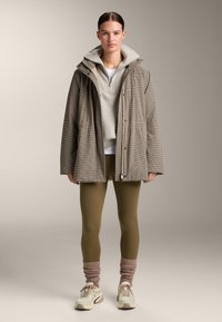 Vrouw draagt een geruite oversized jas, een beige trui met rits, olijfgroene leggings, beige geplooide beenwarmers en witte sneakers, staat stil.
