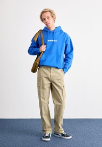 Blå hoodie med vit logotyp, beige cargobyxor och svarta sneakers. Modellen håller en brun väska och står mot en neutral bakgrund.