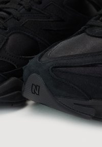Paire de baskets noires avec détails en suède et mesh, semelles en caoutchouc incurvées, et logo "N" embossé sur le côté extérieur près des orteils.