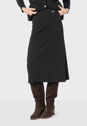 Jupe noire genouillère en tissu doux, avec un design simple et un subtil accent métallique à la taille. Associée à des bottes en daim marron.