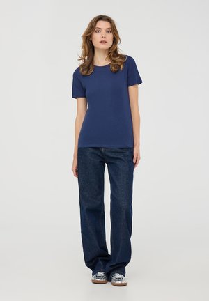 T-shirt di cotone blu navy con maniche corte, abbinato a jeans in denim scuri a gamba larga e scarpe con fantasia. Sfondo semplice.