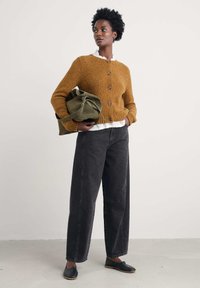 Vrouw staat met krullend haar, een bruine knoptrui, een witte blouse, wijde zwarte spijkerbroek, zwarte schoenen en houdt een olijfgroene tas vast.