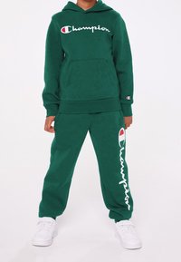 Grön fleecehoodie med en framficka och vit "Champion"-logotyp. Matchande gröna fleecebyxor med logotryck längs benet. Vita sneakers.