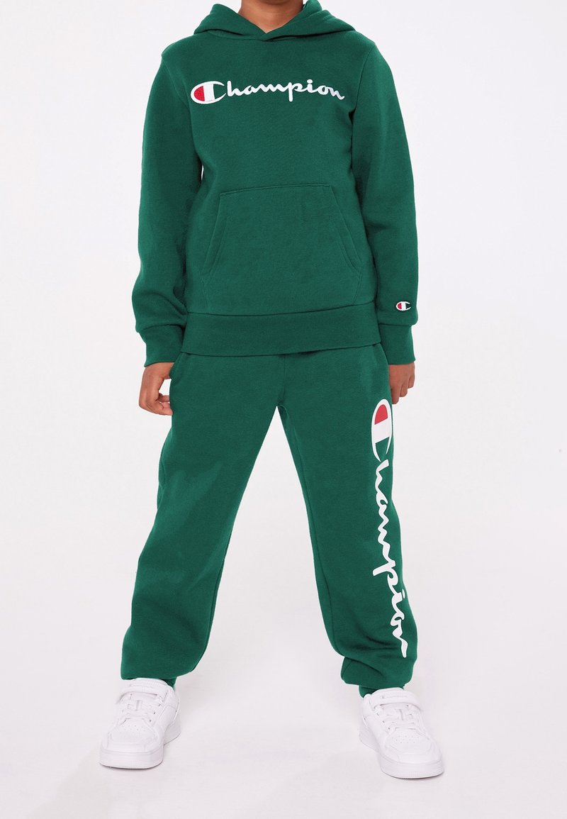 Grön fleecehoodie med en framficka och vit "Champion"-logotyp. Matchande gröna fleecebyxor med logotryck längs benet. Vita sneakers.