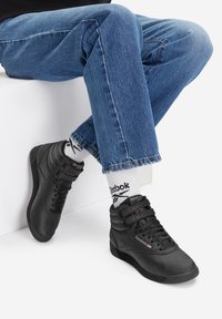 Mustad mustad tennisjalatsid mitme paelaringiga, tekstureeritud nahast ülemine osa, pehme pahkluu tugi ja Reebok'i bränding. Kombineeritud siniste teksade ja valgete sokkidega.