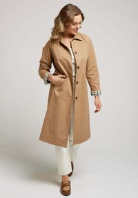 Manteau beige avec un col, des boutons et deux poches latérales. Les poignets présentent un motif à carreaux. Porté par-dessus une tenue de couleur claire.
