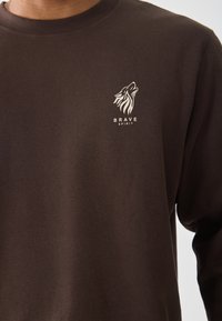 Sudadera de algodón marrón con cuello redondo y un gráfico de lobo con el texto "BRAVE SPIRIT" en blanco en el lado izquierdo del pecho.