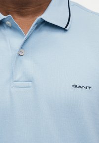 Ανοιχτό μπλε πόλο μπλουζάκι με δύο κουμπιά, σκούρο περίγραμμα στο γιακά και κεντημένο λογότυπο "GANT" στο στήθος.