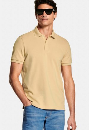 Man met zwarte zonnebril, beige polo met korte mouwen en blauwe spijkerbroek, staand met één hand in de zak tegen een witte achtergrond.