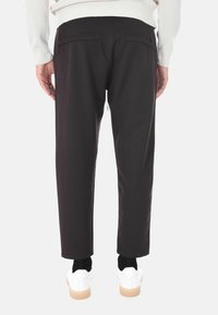 Uomo che indossa pantaloni neri a gamba dritta, sneakers bianche e un maglione bianco a maniche lunghe, mostrato di spalle su uno sfondo bianco.