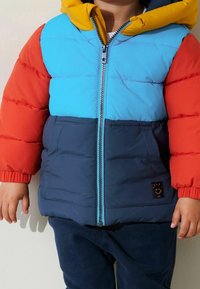Giacca imbottita con un design a blocchi di colore che presenta sezioni blu, rosse e gialle, chiusura con zip e una piccola toppa con logo sul lato inferiore.