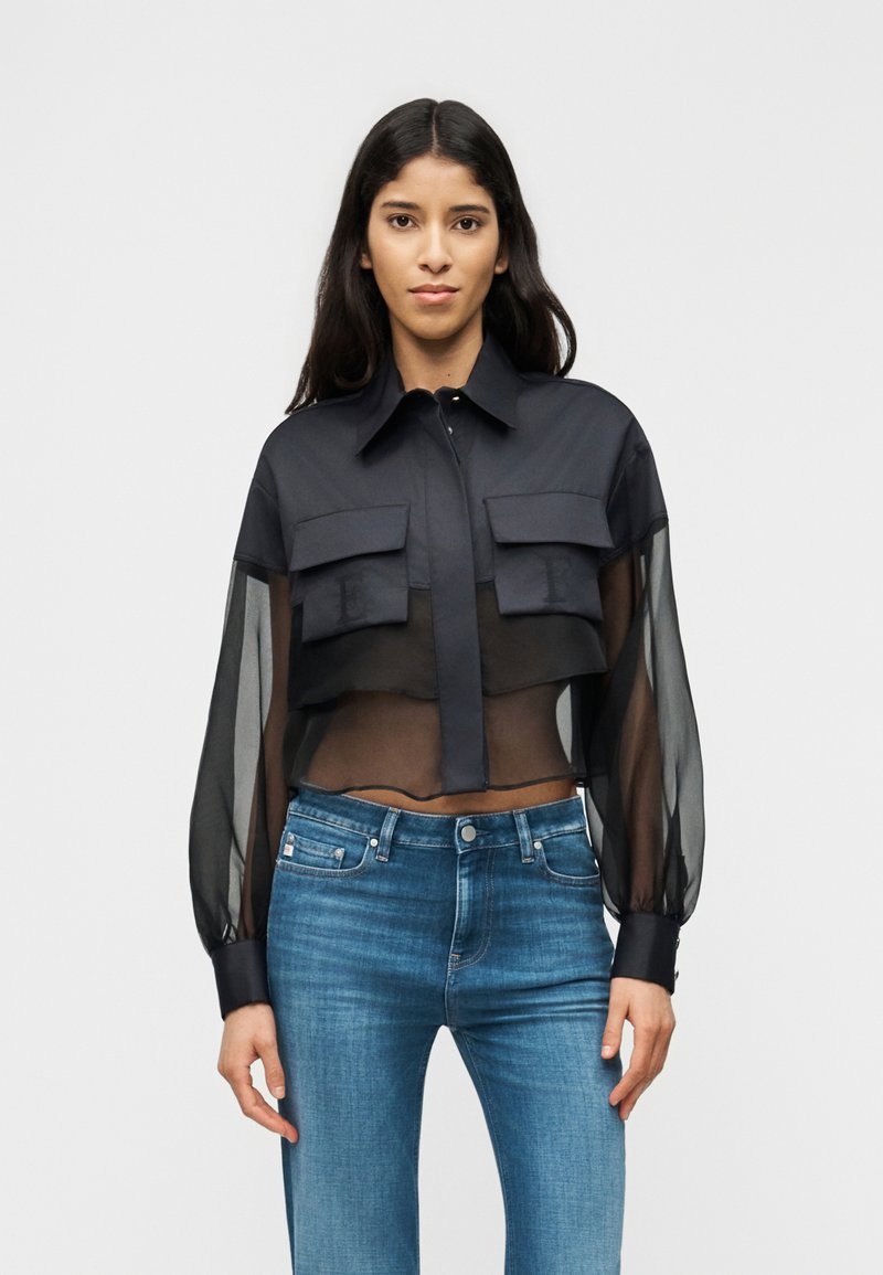 Γυναίκα που φοράει μαύρο διαφανές κοντό μπλούζα με μακριά μανίκια και μπλε ψηλόμεσο τζιν, στέκεται μπροστά σε ένα απλό φωτεινό φόντο.