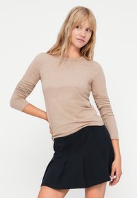 Beżowa bluzka z długim rękawem wykonana z miękkiego materiału, z okrągłym dekoltem i dopasowanym krojem, zestawiona z czarną plisowaną spódnicą.