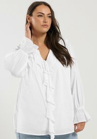 White blouse with ruffled front detail, v-neckline, and long puffed sleeves. Made of light fabric with a relaxed fit and subtle texture.