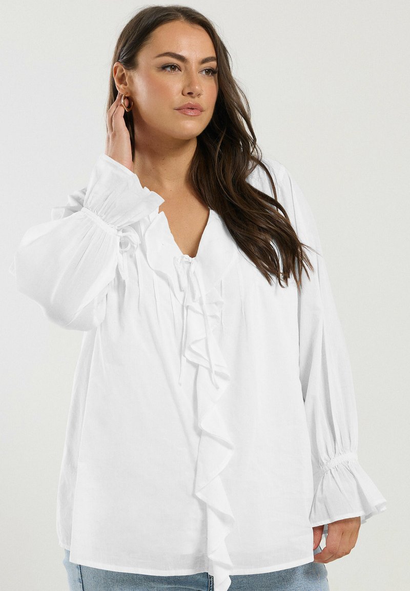 White blouse with ruffled front detail, v-neckline, and long puffed sleeves. Made of light fabric with a relaxed fit and subtle texture.