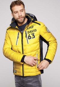 Żółta kurtka puchowa z czarnymi akcentami, z zamkiem błyskawicznym z przodu, dwoma zamykanymi kieszeniami i nadrukowanymi detalami na piersi oraz rękawie.