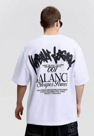 Osoba nosi bijelu široku majicu s crnim tekstom u stilu grafita i izrazima o ravnoteži i snazi na leđima, stoji ispred jednobojne pozadine.