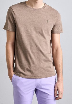 Mand iført en simpel brun t-shirt med korte ærmer og lyse lilla bukser, stående med den ene hånd i lommen mod en grå baggrund.