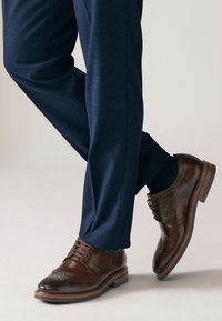 Scarpe brogue in pelle marrone con finitura lucida, caratterizzate da dettagli traforati e suola in gomma, abbinate a pantaloni blu navy.