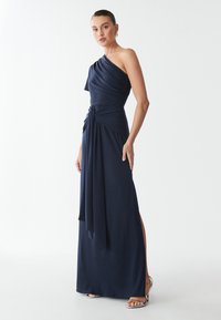 Abito blu navy a spalla unica con tessuto drappeggiato, dettaglio a nodo in vita e un'alta spaccatura su un lato. Texture morbida e fluida.