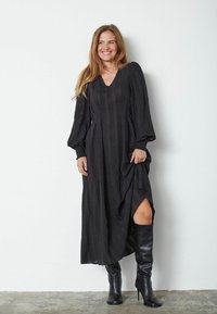 Čierna maxi šaty s výstrihom do V, dlhými rukávmi, textúrovanou tkaninou a bočným rozparkom. Skombinované s čiernymi holými podpätkami po kolená.