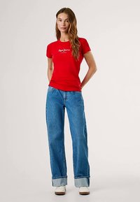 Camiseta roja de algodón con cuello redondo y mangas cortas, con estampado de logo blanco. Combinada con vaqueros azules de pierna ancha y zapatillas blancas.